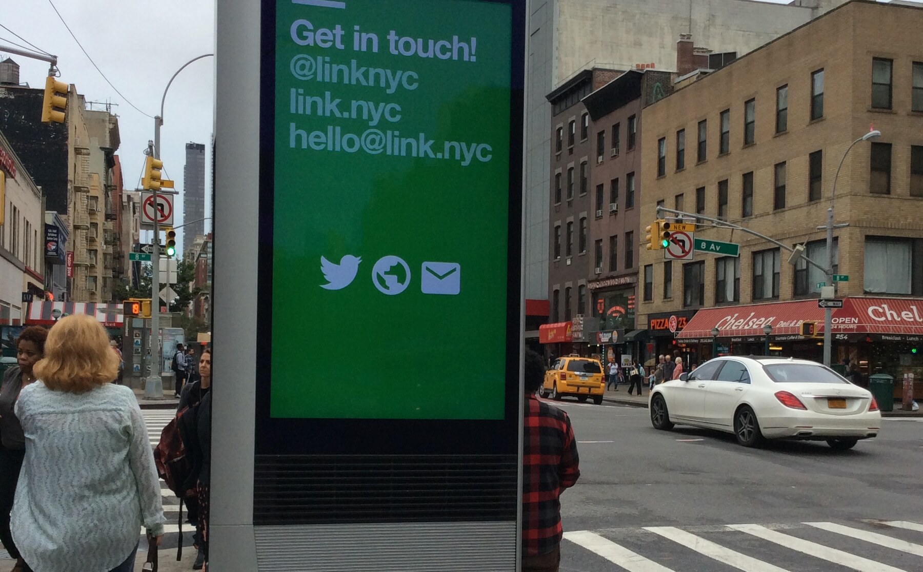 A LinkNYC Link 1.0 kiosk at 23rd Street and 8th Avenue in Chelsea, Manhattan, displaying a green 'Get in touch!' screen with pedestrians and Chelsea storefronts.