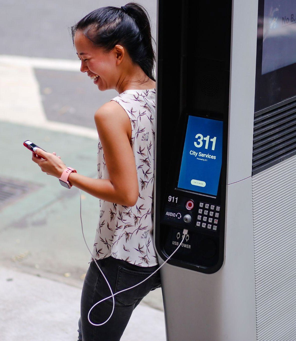 A New Yorker smiling while charging her phone at a LinkNYC kiosk displaying the 311 City Services screen.